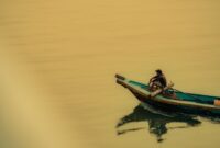 Seorang nelayan tradisional menggunakan perahu kayu memancing ikan dengan metode ramah lingkungan di perairan pesisir Indonesia saat matahari terbenam. Praktik penangkapan ikan secara tradisional ini membantu menjaga kelestarian terumbu karang serta populasi biota laut Nusantara.