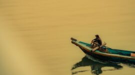 Seorang nelayan tradisional menggunakan perahu kayu memancing ikan dengan metode ramah lingkungan di perairan pesisir Indonesia saat matahari terbenam. Praktik penangkapan ikan secara tradisional ini membantu menjaga kelestarian terumbu karang serta populasi biota laut Nusantara.